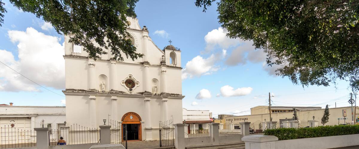 A white church with a clock tower and a cross on top, surrounded by a paved area and greenery.