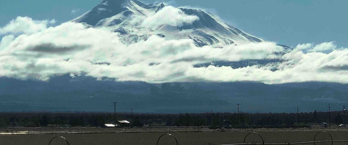 A snow-capped mountain with clouds below, a clear sky, and a road with barbed wire in the foreground.