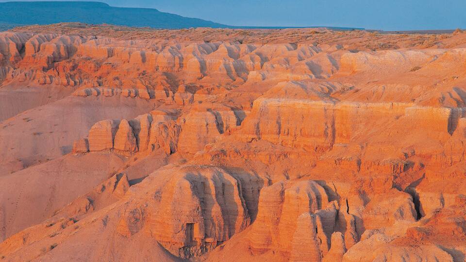 A vast desert landscape with orange rock formations and a distant mountain range.