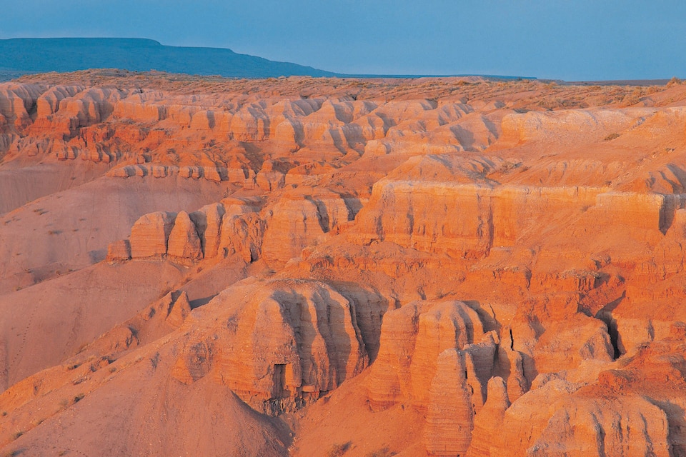 A vast desert landscape with orange rock formations and a distant mountain range.