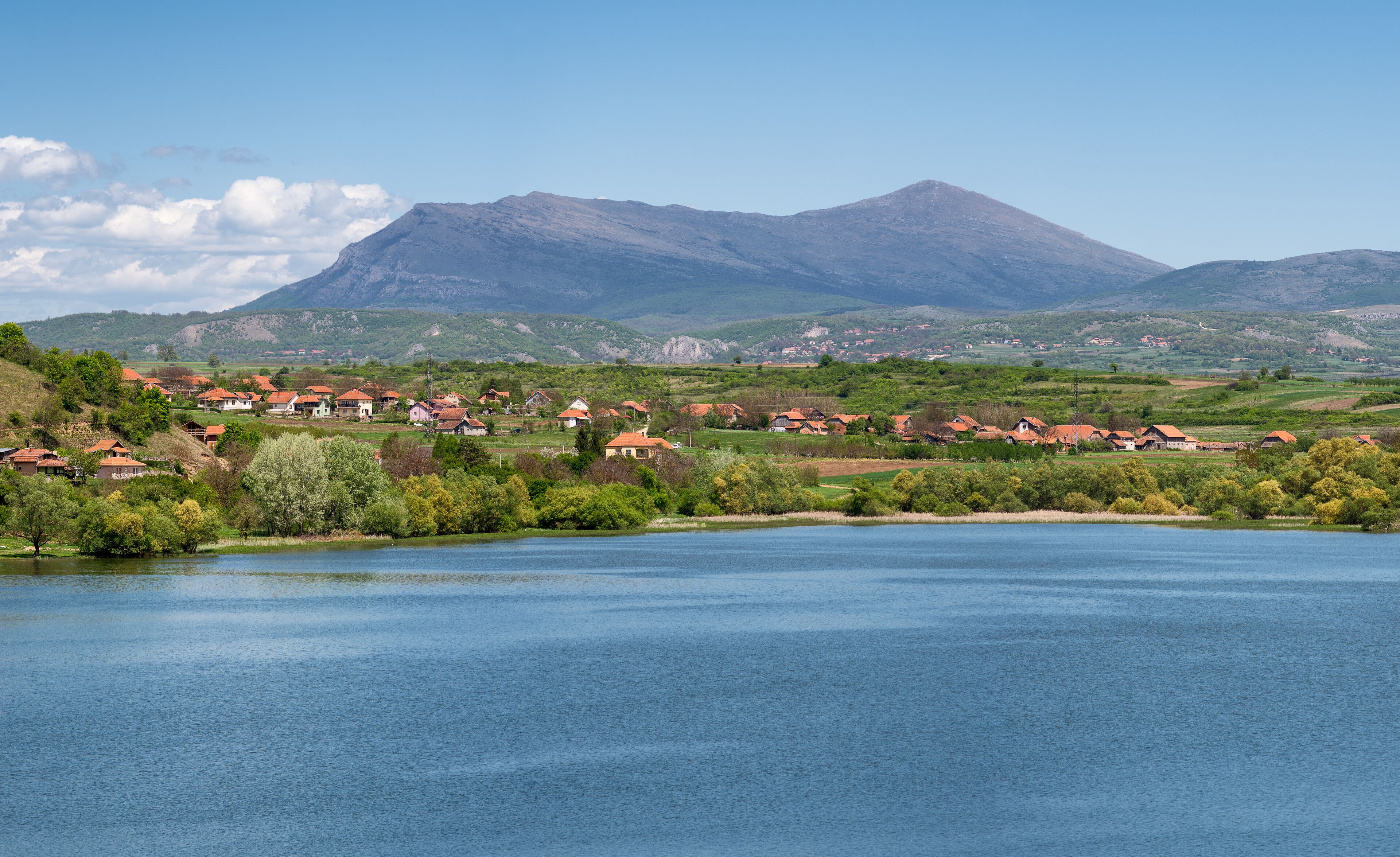 A lakeside village with red-roofed houses, surrounded by green hills and a mountain in the background.