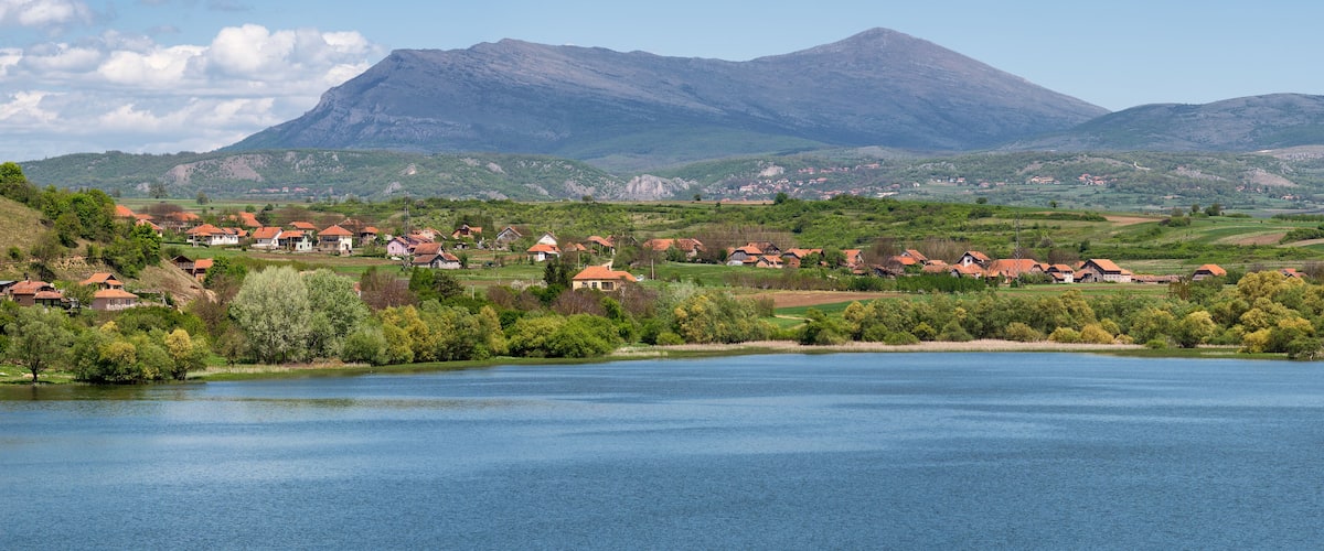 A lakeside village with red-roofed houses, surrounded by green hills and a mountain in the background.