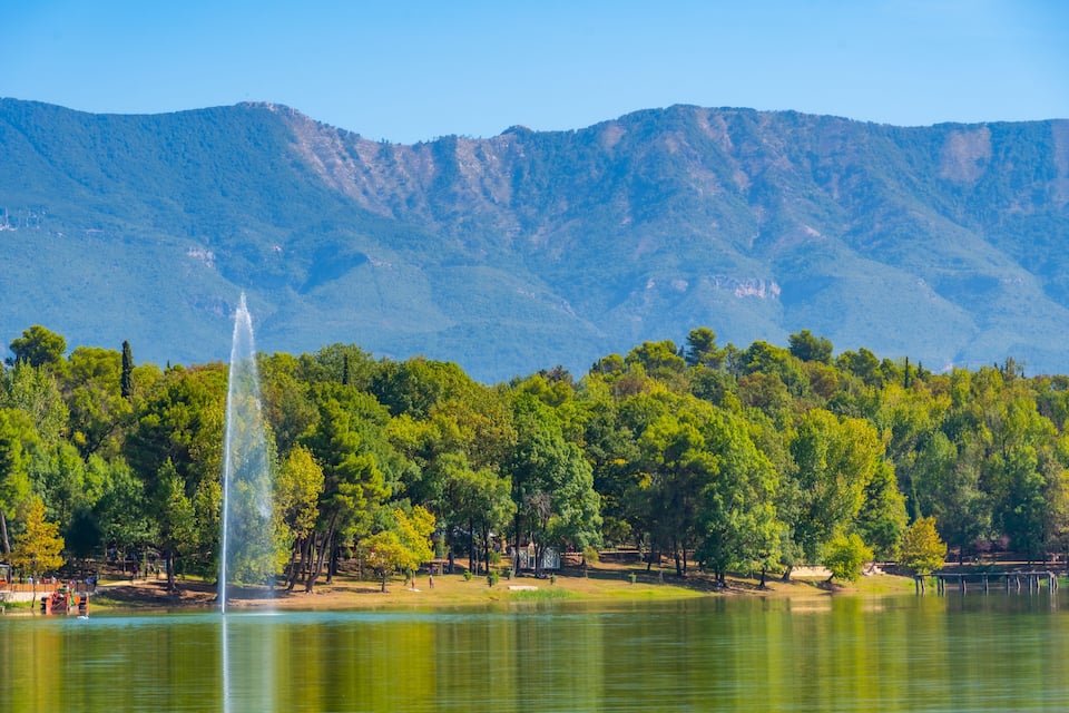 Un lago con una fuente, rodeado de árboles verdes y densos y montañas al fondo.
