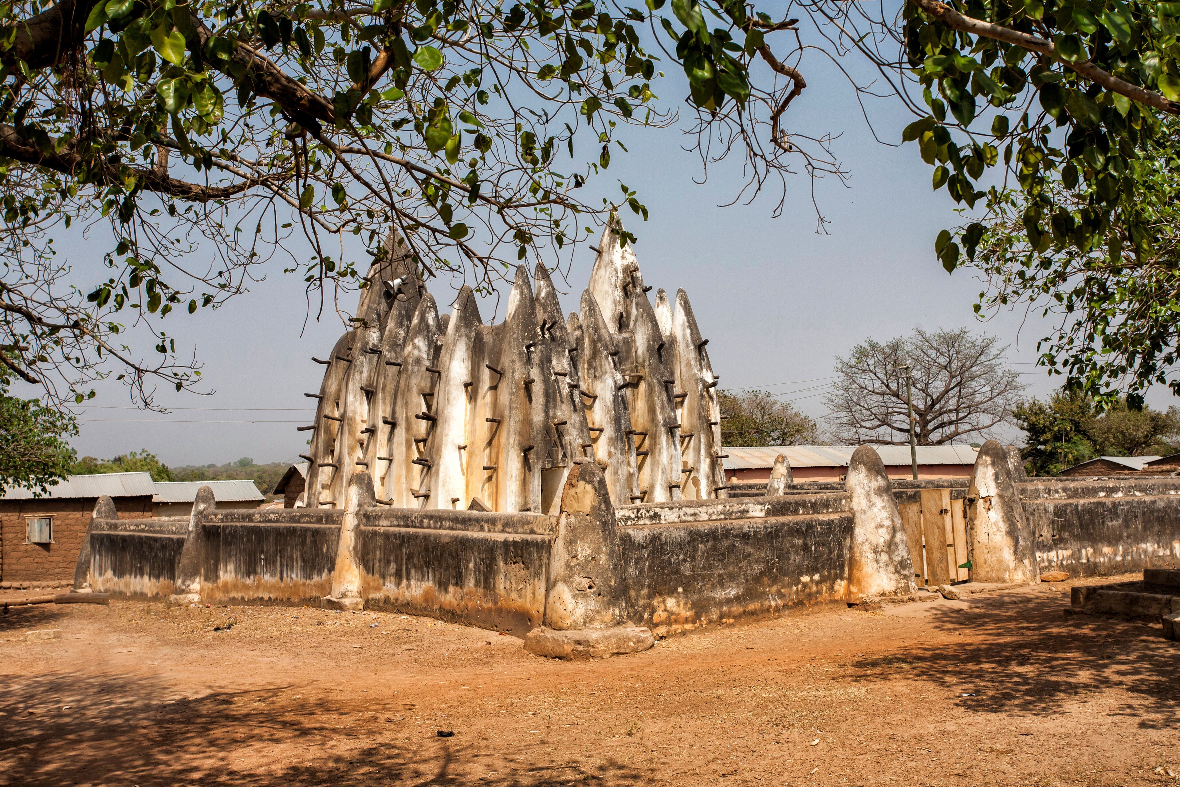 A historical stone wall with pointed tops, surrounded by a dry, open landscape with sparse vegetation and a few buildings in the background.