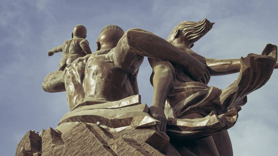 A bronze statue of a seated figure with a child on its back, set against a clear sky.