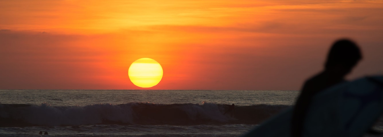 Un atardecer sobre el océano con la silueta de una persona sosteniendo una tabla de surf.