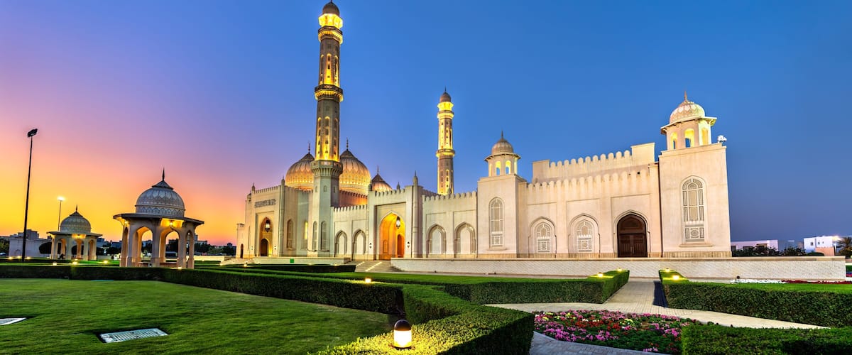 The beautiful Sultan Taimur bin Faisal Mosque in Seeb, Oman, illuminated during a colorful sunset, showcasing intricate Islamic architecture and landscaped gardens.
