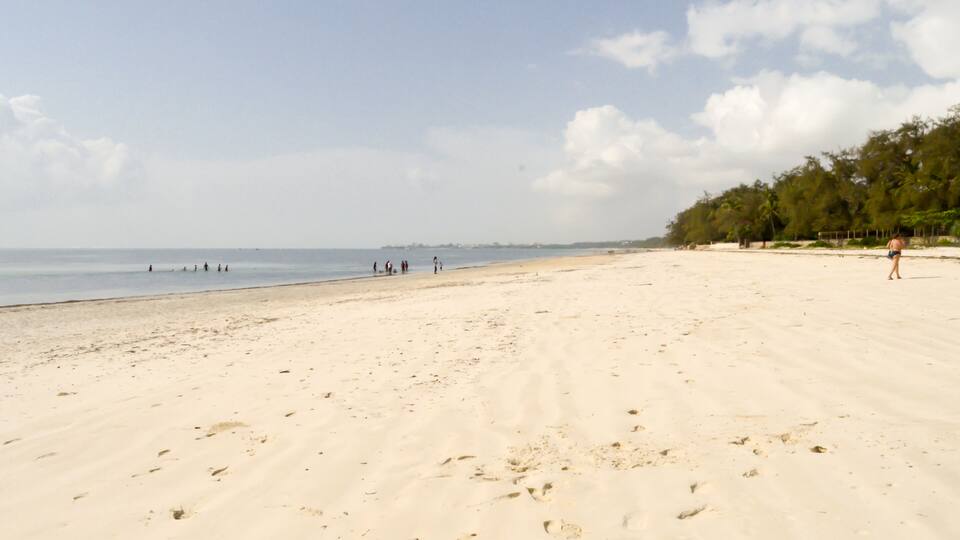 A wide sandy beach with footprints, a calm sea, and a line of trees in the background.