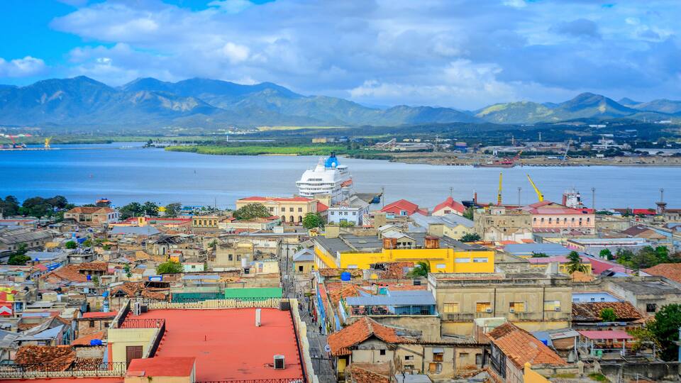A coastal city with a prominent white dome structure, colorful buildings, and mountains in the background.