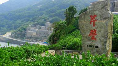 A stone with Chinese characters and the text "CHIN BE" in English, surrounded by lush greenery and a coastal village in the background.