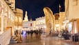A nighttime street scene with a large golden spiral sculpture in the center, surrounded by traditional buildings and a mosque with a minaret.