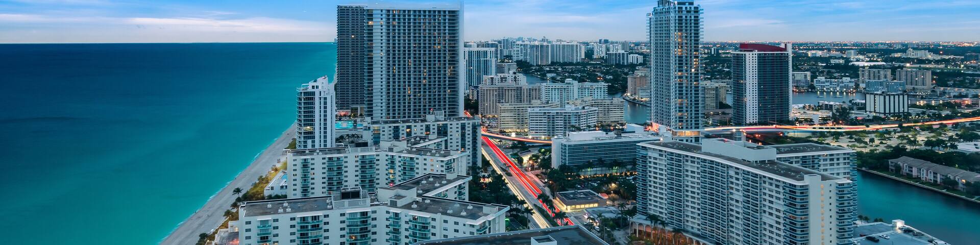 A coastal cityscape with high-rise buildings, a beach, and a busy highway with light trails.