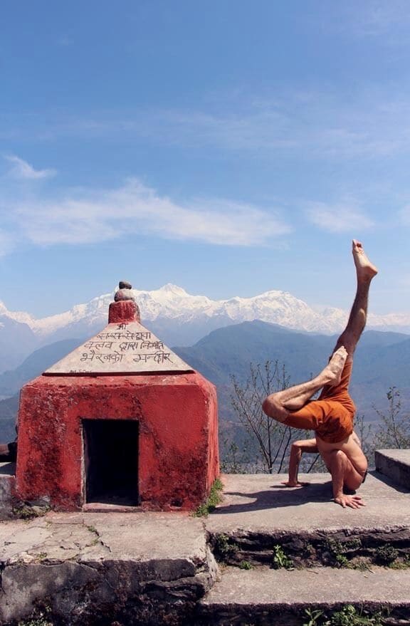 A person performing a handstand on a concrete platform with a red structure in the foreground and snow-capped mountains in the background.