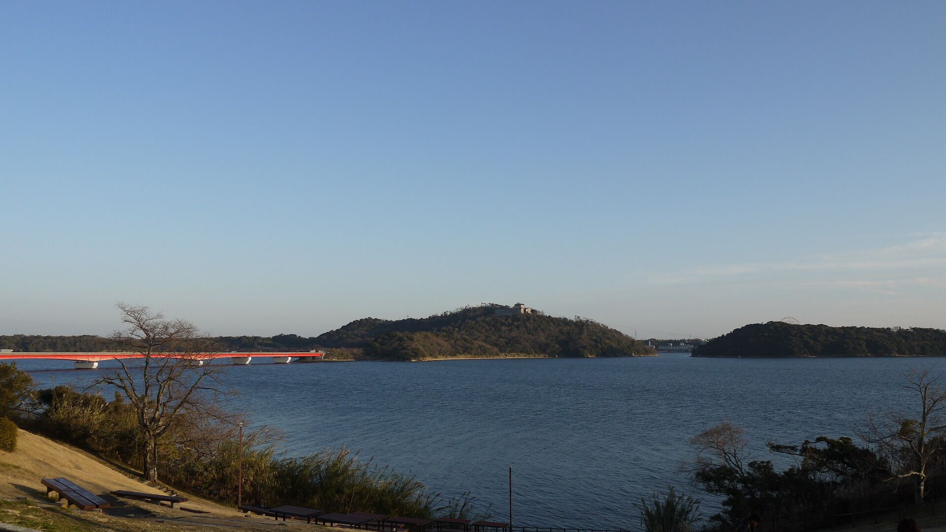 A river with a red bridge, a hill with a building, and picnic tables along the shore.