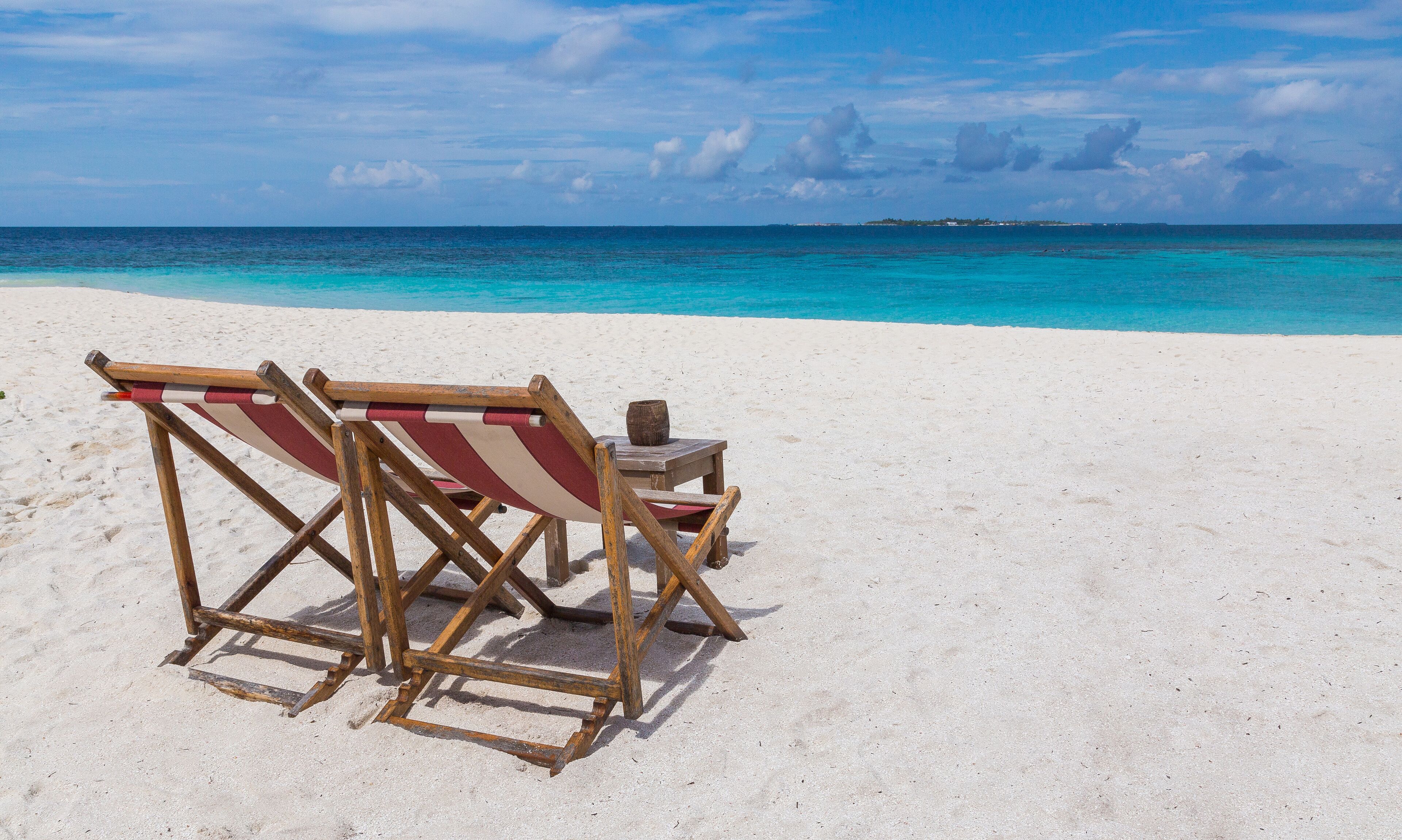 A beach with a wooden lounge chair and a small table with a vase.