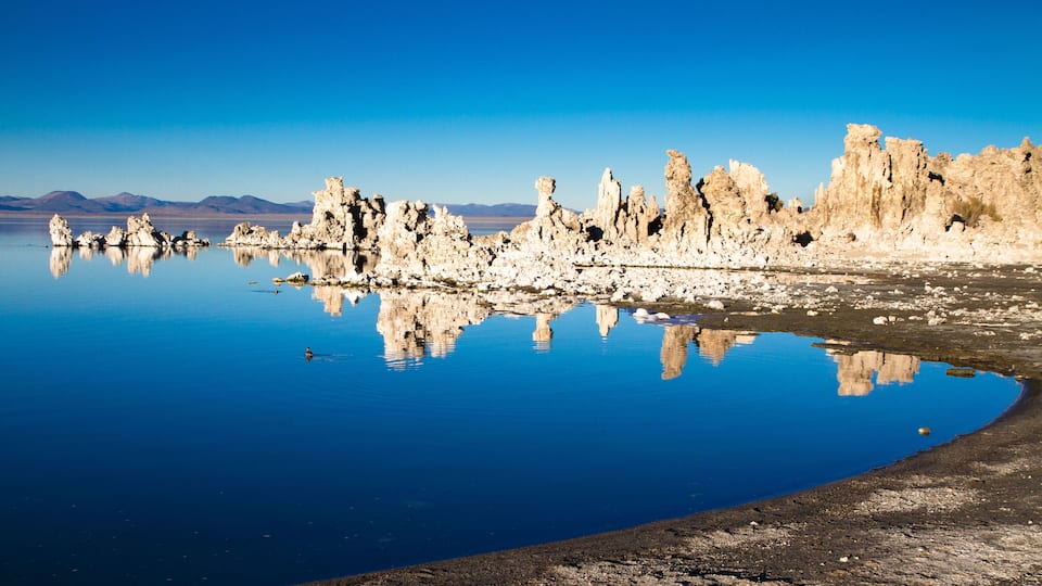 A calm lake with unique rock formations reflecting in the water, surrounded by a clear blue sky.