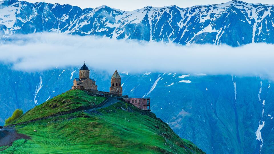 A mountain with snow-capped peaks, a church with two towers, and a lush green hill.