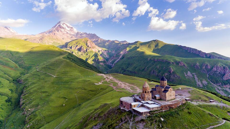 A mountainous landscape with a historic stone building featuring a bell tower, surrounded by lush green valleys and a prominent mountain peak in the background.