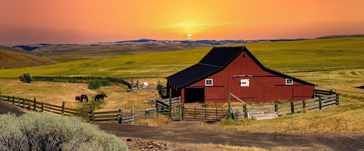 A red barn with a black roof, surrounded by a wooden fence, set against a sunset over rolling hills.