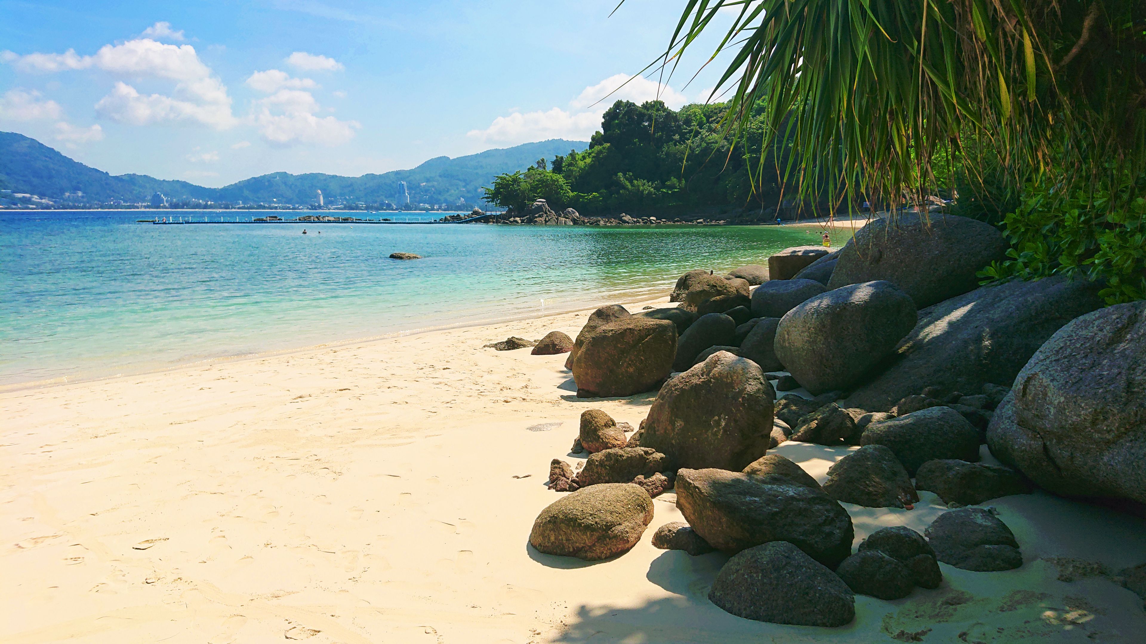 A beach with large rocks, clear turquoise water, and a distant mountain range.