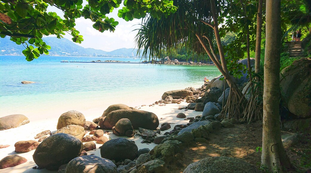 A tropical beach with large rocks, clear turquoise water, and a distant mountain range.