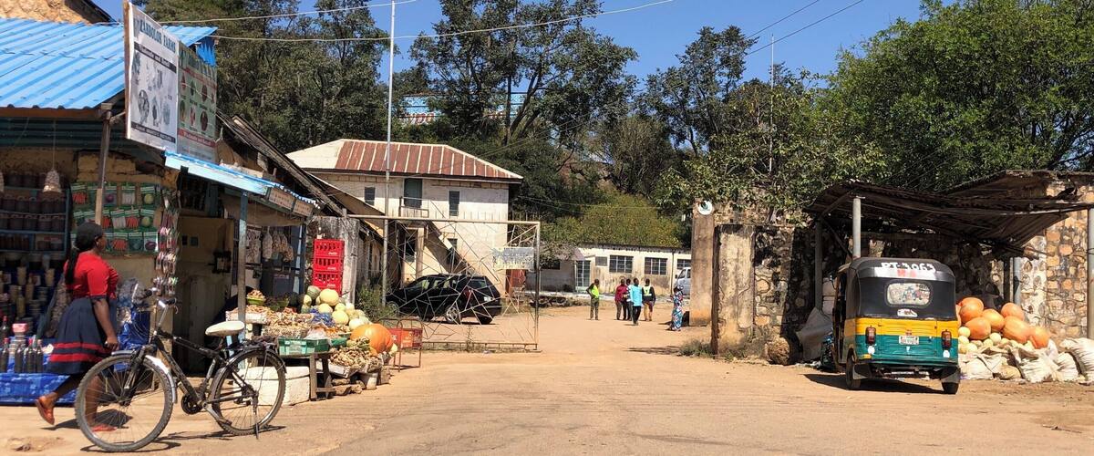 A rural market scene with a dirt road, a bicycle, a three-wheeled vehicle, and a group of people.