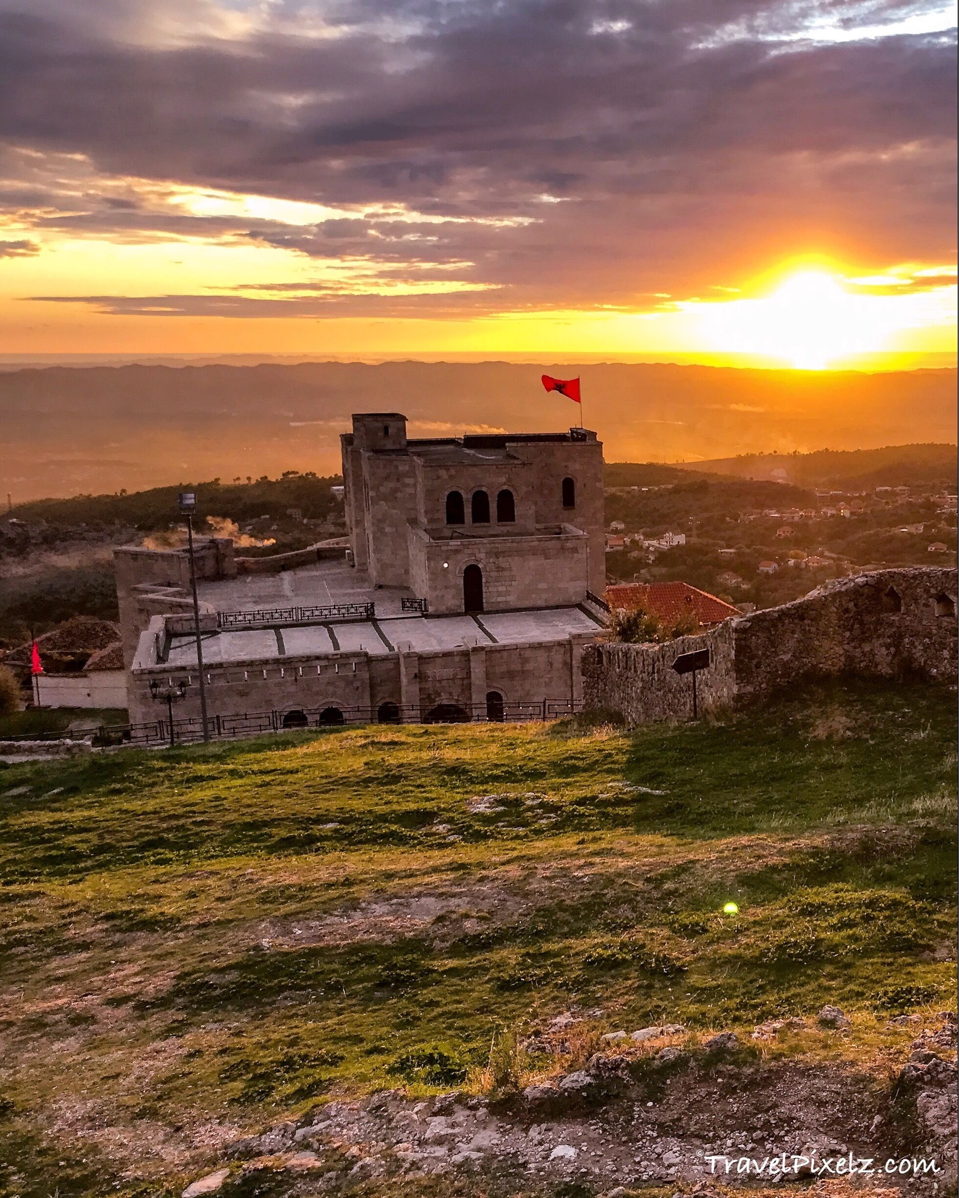 A historic stone building with a red flag, surrounded by a hilly landscape and a sunset in the background.