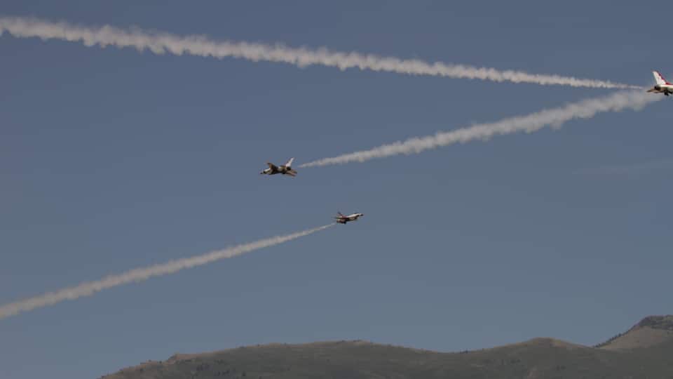 Three jets flying in formation with smoke trails against a clear blue sky and a hilly landscape below.