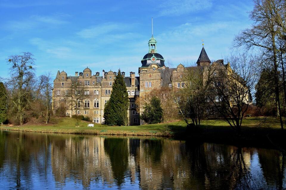 A large castle with a green dome and a spire, surrounded by trees and a lake.