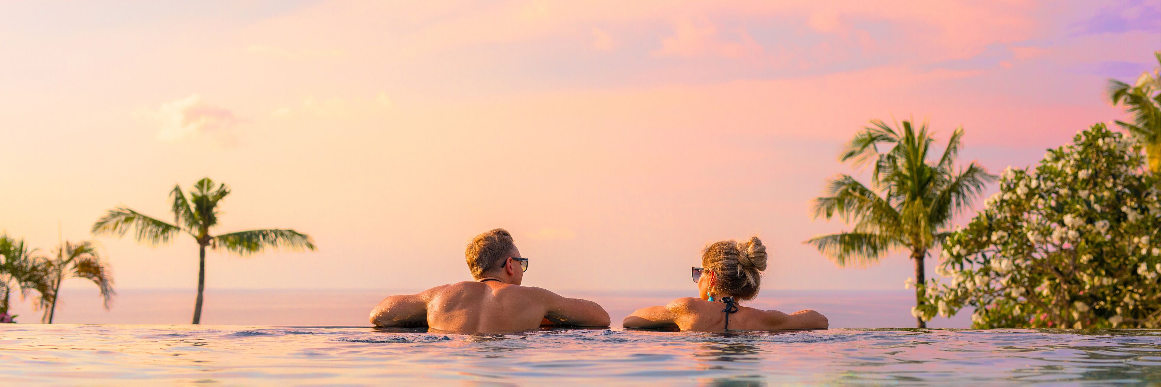 Two people relaxing in a pool with palm trees and a sunset in the background.