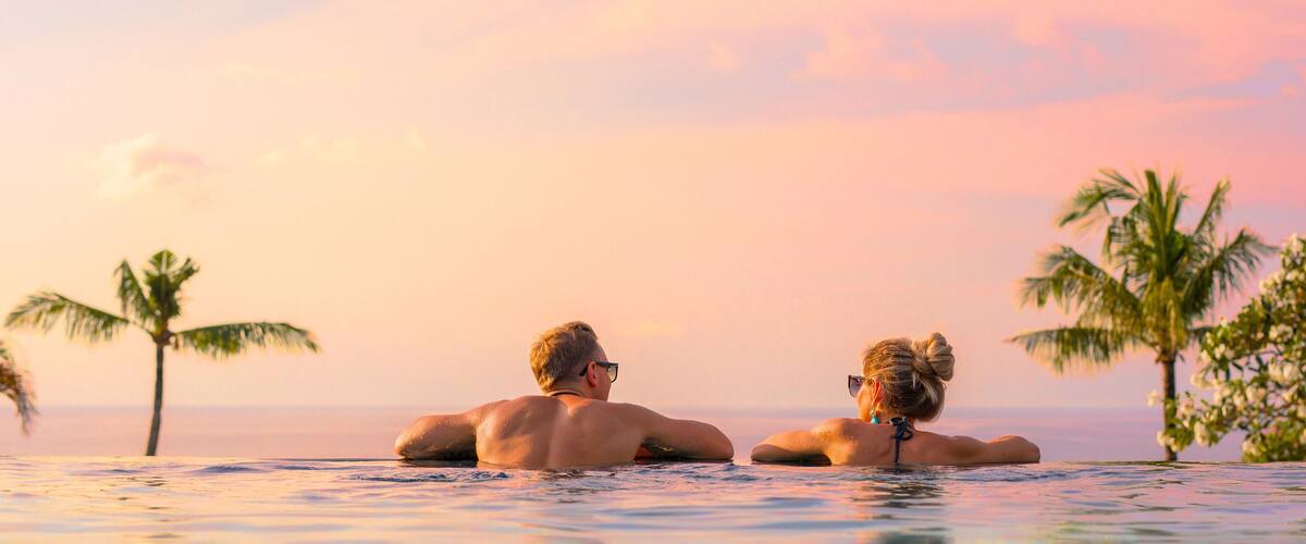 Two people relaxing in a pool with palm trees and a sunset in the background.