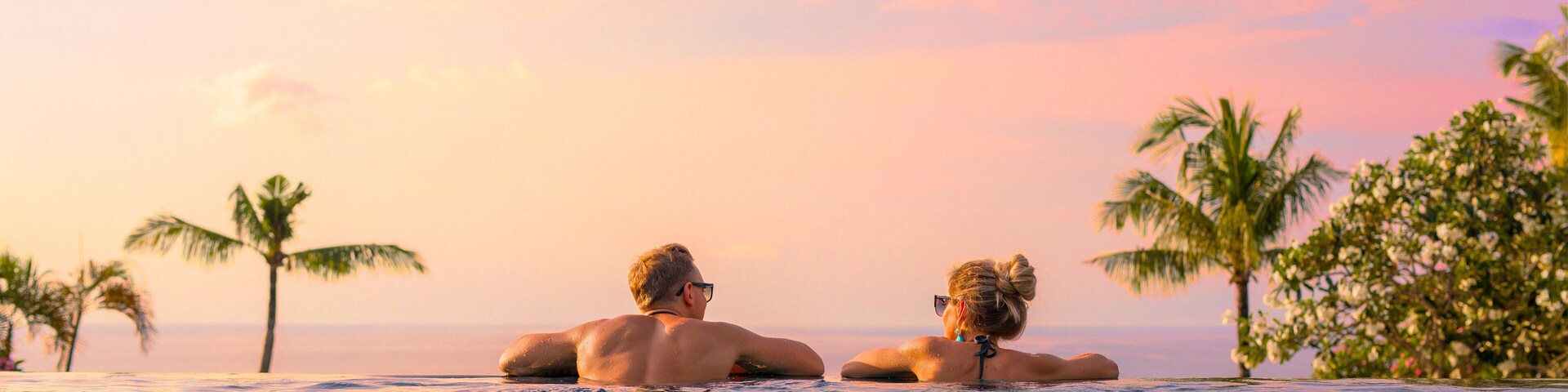 Two people relaxing in a pool with palm trees and a sunset in the background.