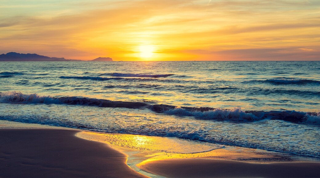 A beach with waves at sunset, a distant mountain, and a clear sky.