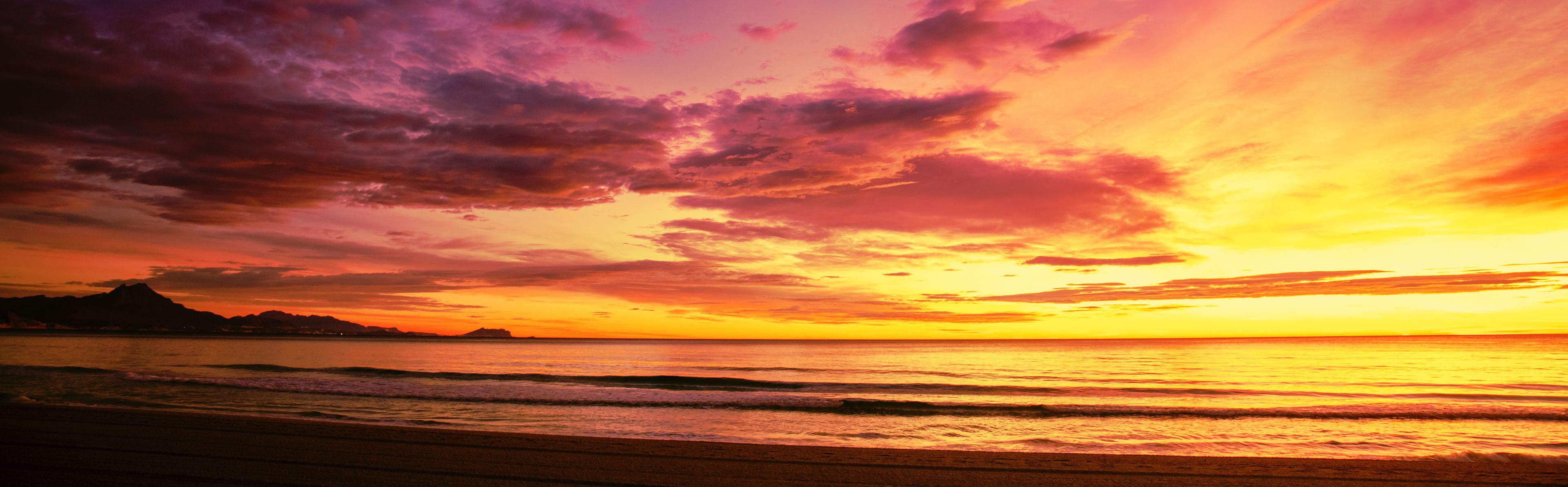 A sunset over a beach with waves, a distant mountain, and a colorful sky.
