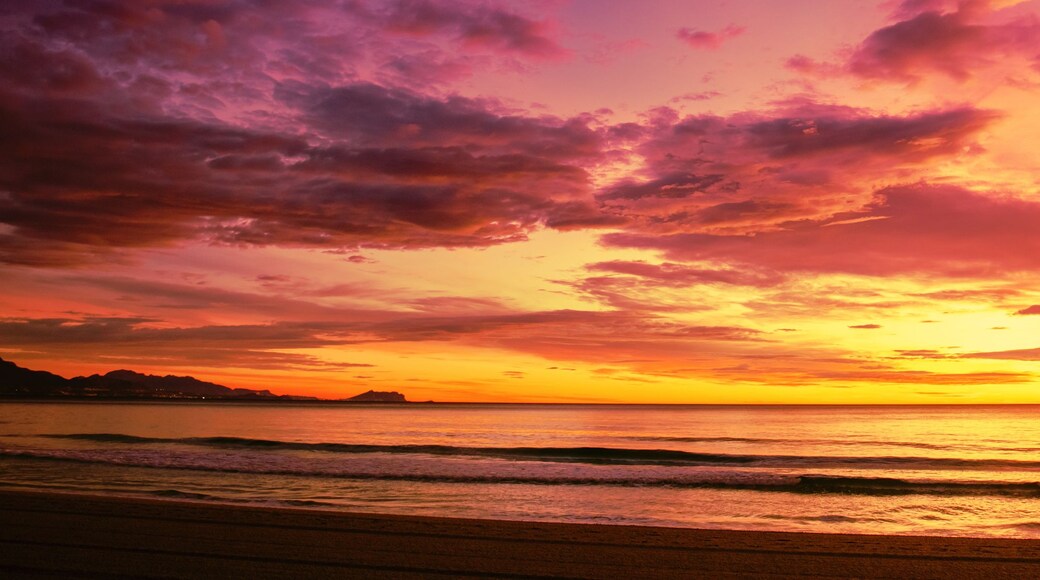 A sunset over a beach with waves, a distant mountain, and a colorful sky.