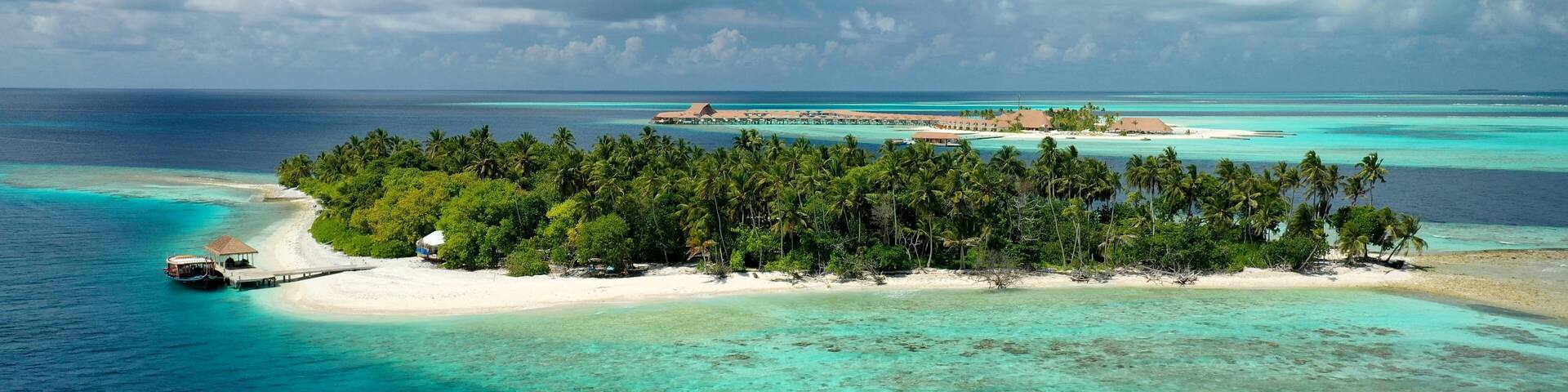 A tropical island with a white sandy beach, dense palm trees, clear turquoise waters, and a small boat docked on the shore.