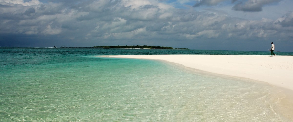 A beach with clear turquoise water, a person walking on the sand, and a distant island.