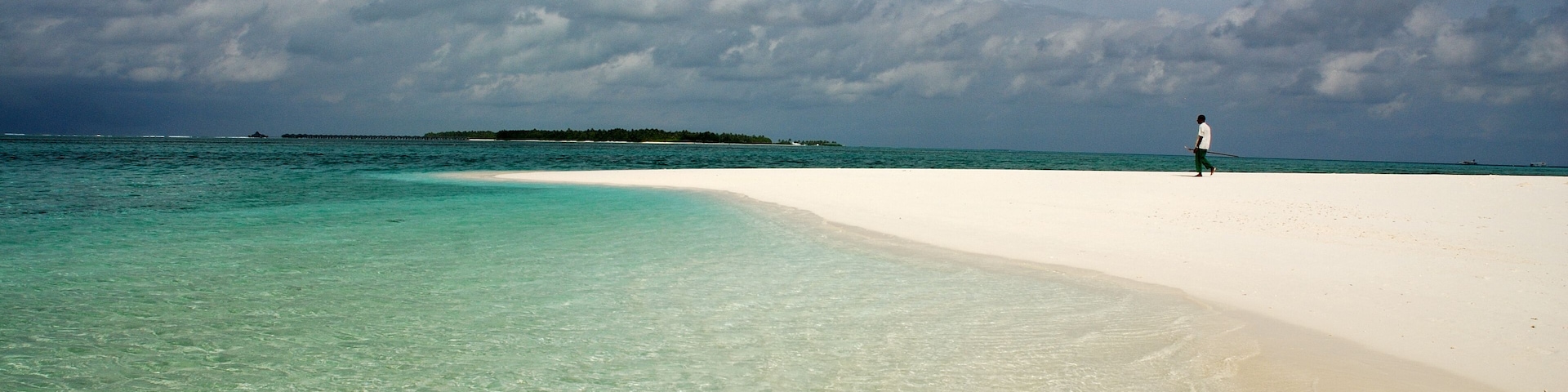 A beach with clear turquoise water, a person walking on the sand, and a distant island.