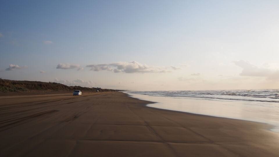 A beach with a sandy shore, a few cars parked, and a clear sky.