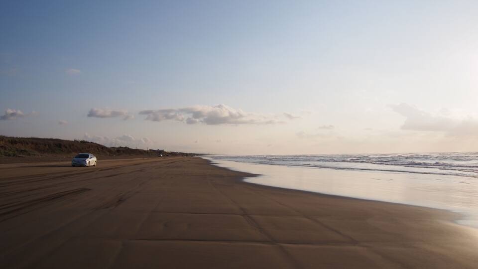 A beach with a car parked on the sand and waves approaching the shore.
