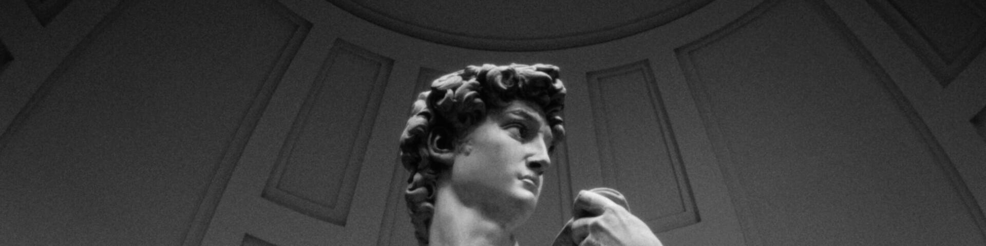 A classical statue of a male figure in a standing pose, wearing a draped garment, set against a backdrop of a curved architectural ceiling with paneling.