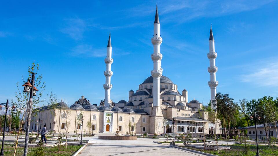 A mosque with three minarets and a large dome, surrounded by a plaza with trees and a clear blue sky.