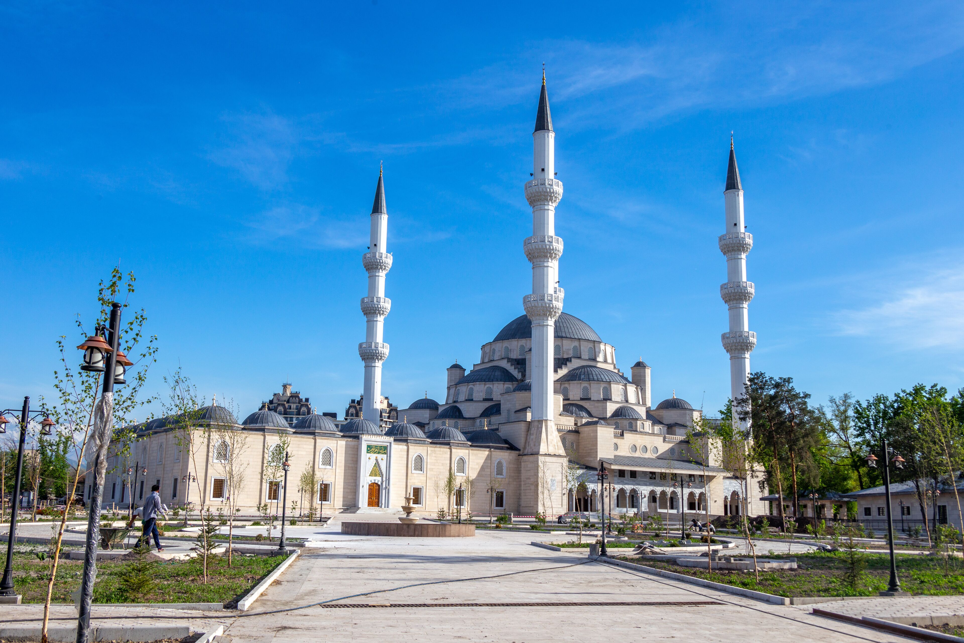 A mosque with three minarets and a large dome, surrounded by a plaza with trees and a clear blue sky.