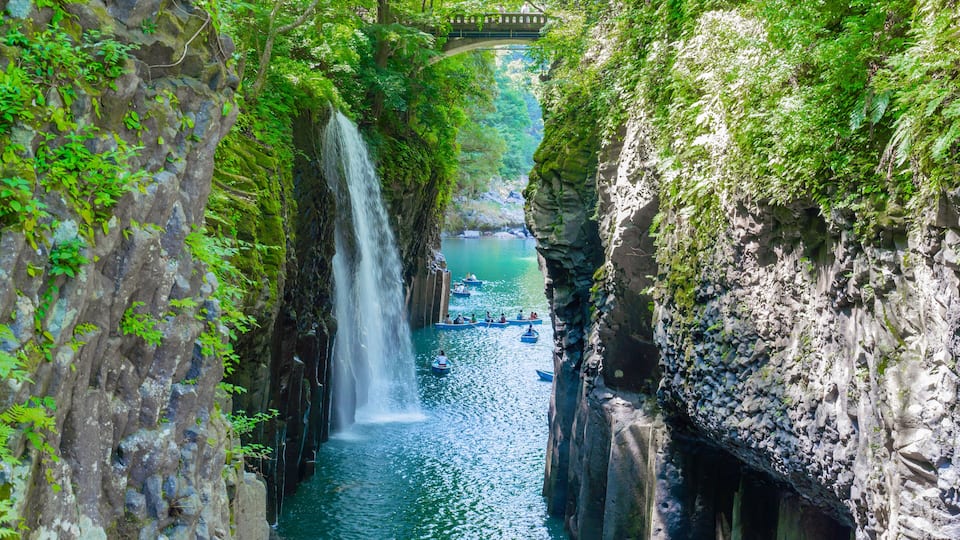 A waterfall cascading down a rocky gorge, surrounded by lush greenery and a bridge in the background.