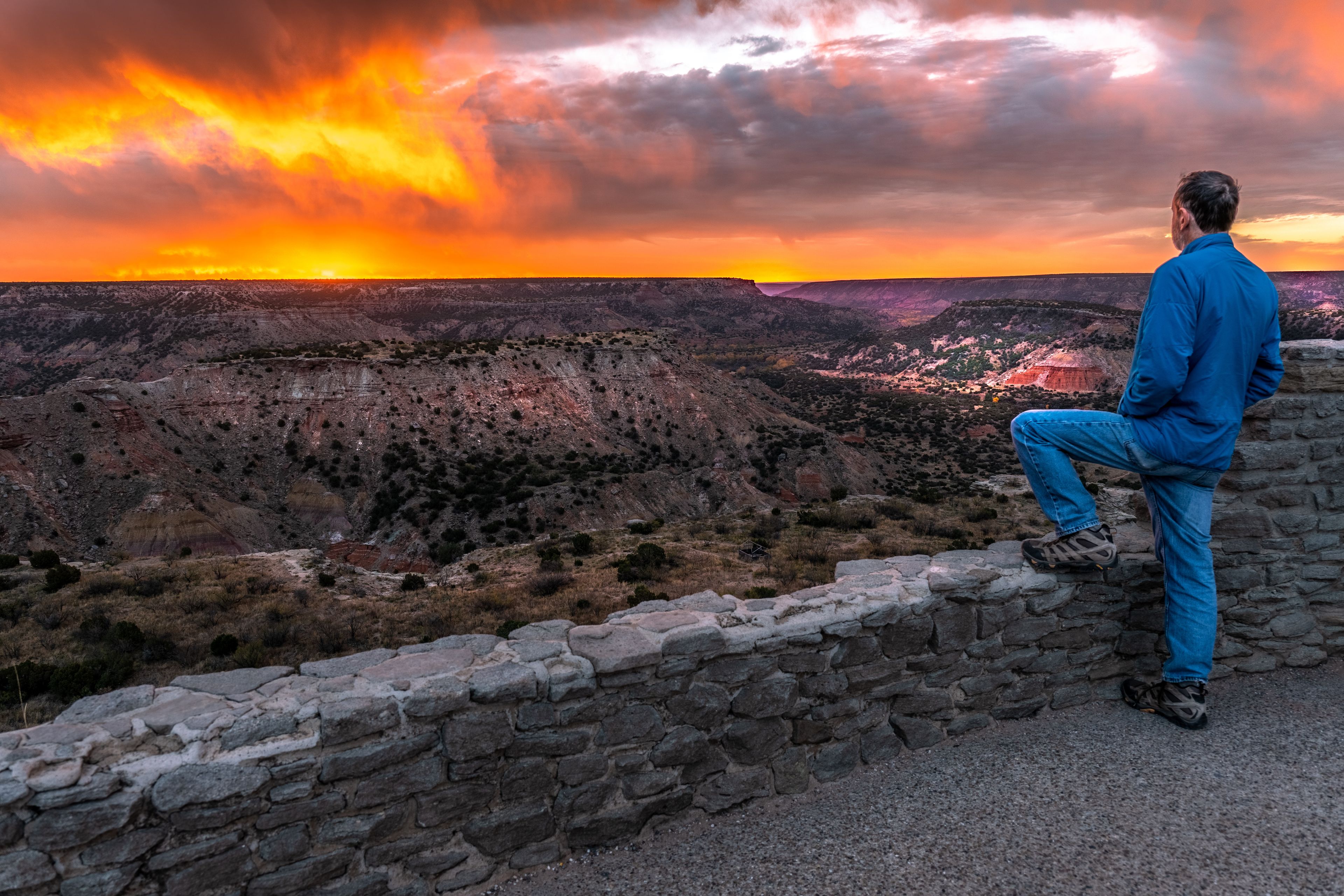A mature man person Caucasian standing on a paved area one leg on stone fence watching a scene of deep arid valley illuminated by bright orange sunrise, Palo Duro Canyon State Park, Texas
