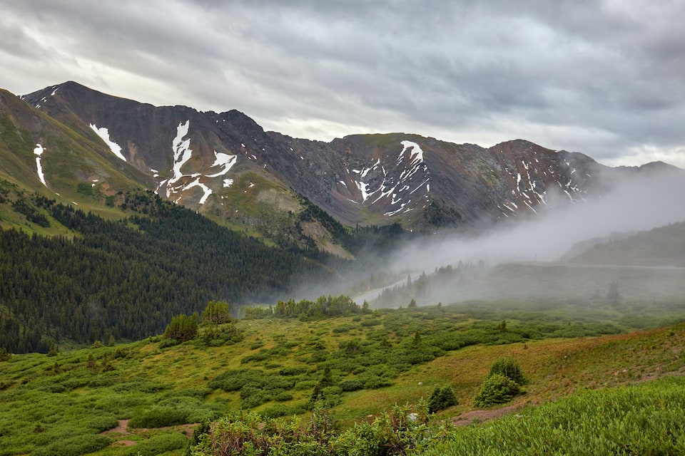 Paisatge de muntanya amb cims nevats, una vall amb boscos densos i una atmosfera brumosa.