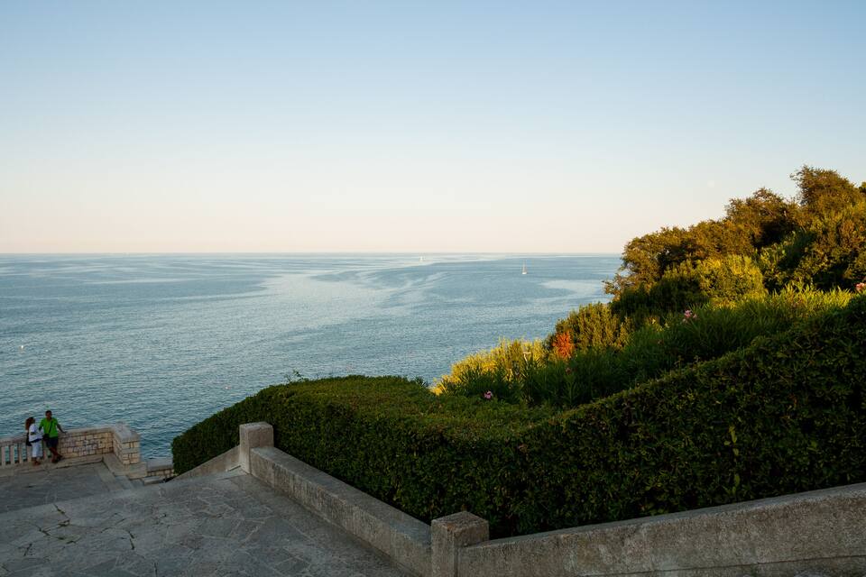 A coastal landscape with a stone staircase leading down to the sea, surrounded by lush greenery and a clear sky.