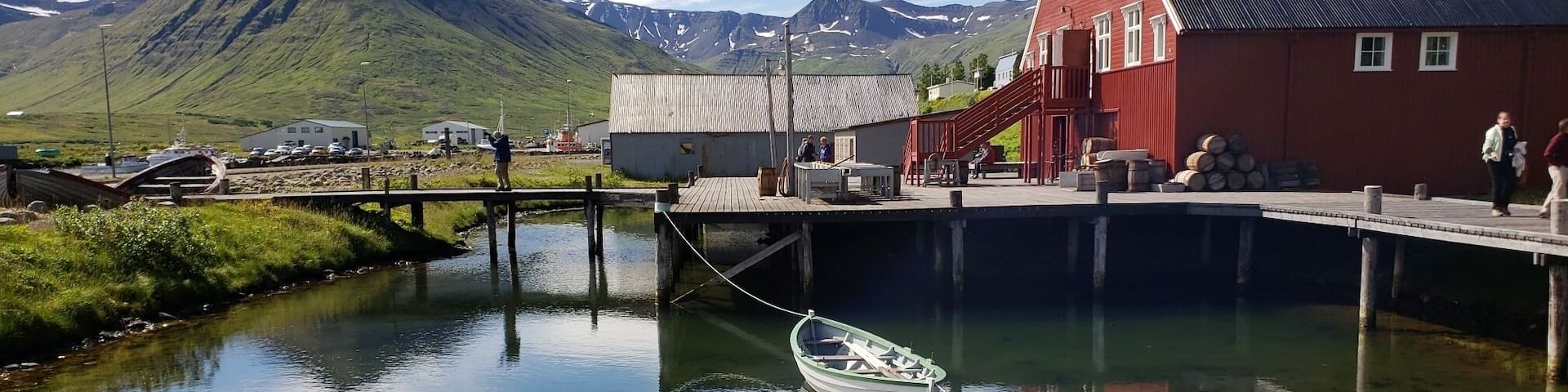 A calm river with a small boat tied to a dock, a red building, and a mountain in the background.