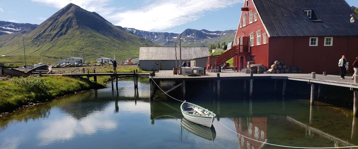 A calm river with a small boat tied to a dock, a red building, and a mountain in the background.