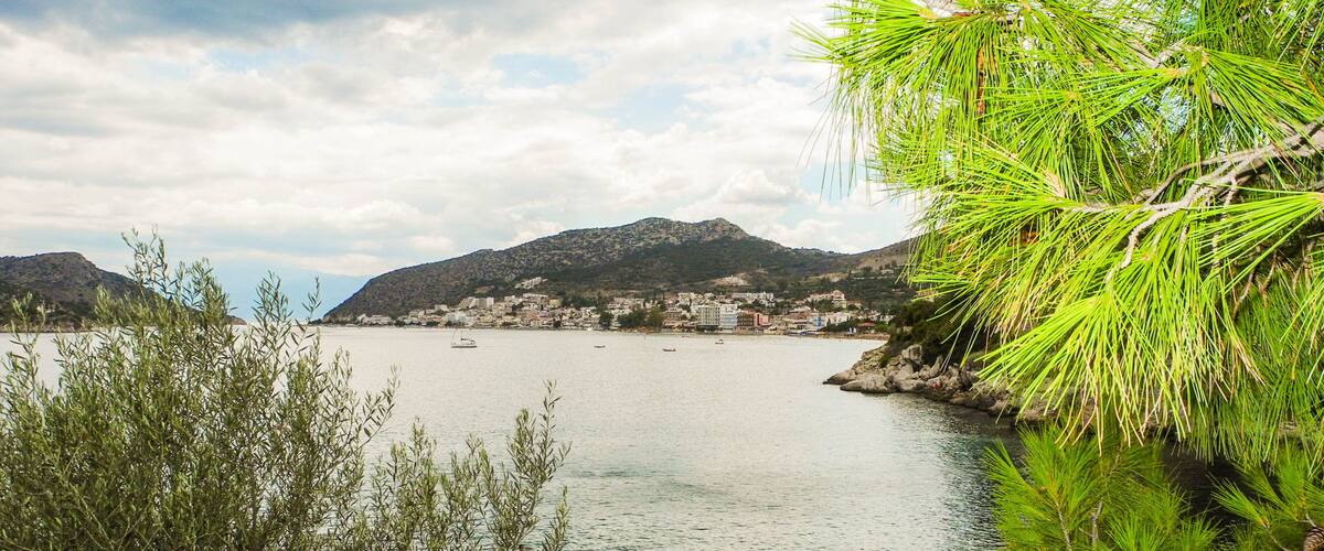 A coastal town with a calm sea, a sailboat, and a mountain in the background.